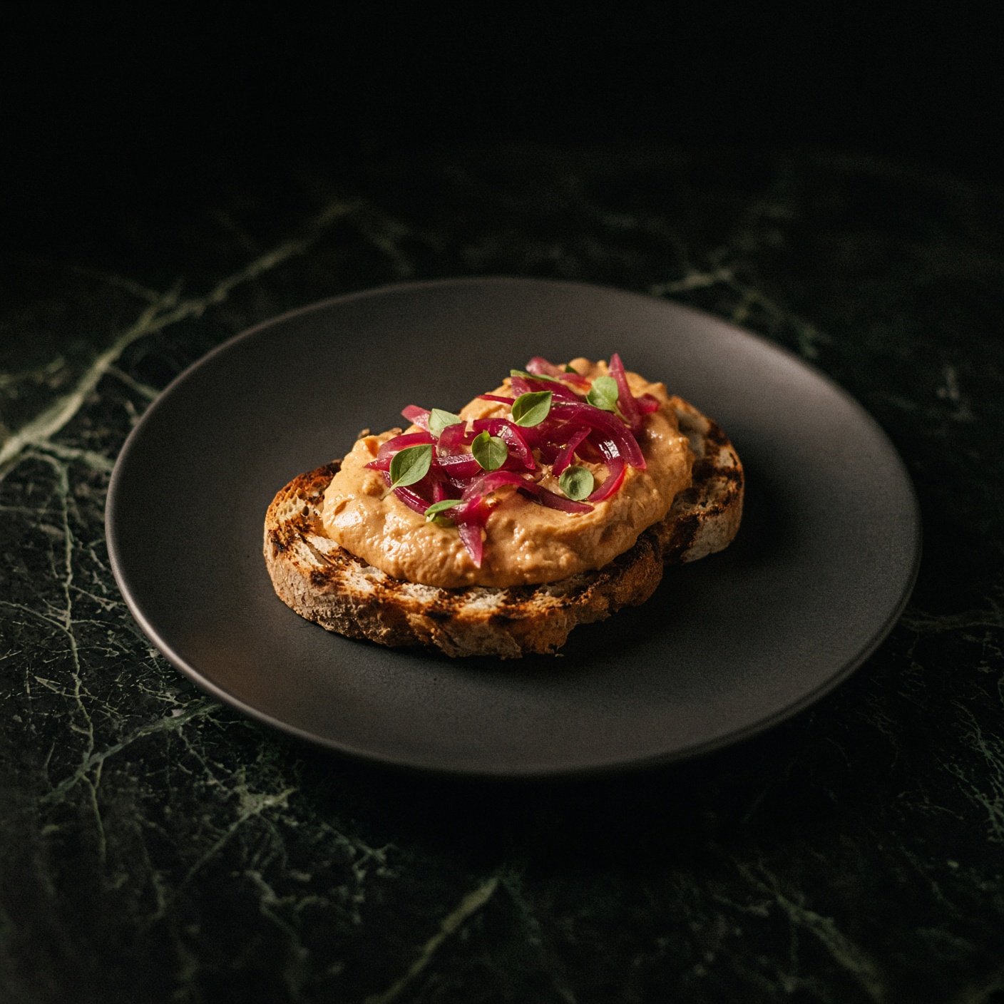 Duck liver parfait on grilled sourdough with pickled red onion and marjoram, on a dark plate.